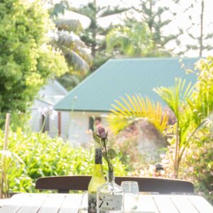 Sunlit garden patio with a wooden table, cushioned chairs, and a single flower in a bottle as a centrepiece.
