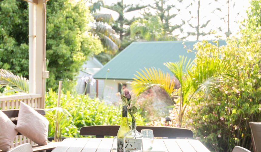 Sunlit garden patio with a wooden table, cushioned chairs, and a single flower in a bottle as a centrepiece.
