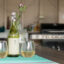 Two glasses of white wine beside a bottle on an outdoor table with a barbecue grill in the background.