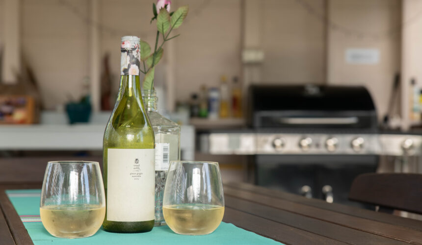 Two glasses of white wine beside a bottle on an outdoor table with a barbecue grill in the background.